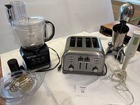 View of all three appliances together including the Black & Decker food processor with blades and lid, Cuisinart stainless steel toaster, and Braun immersion blender with mixing cup and milkshake maker attachment.