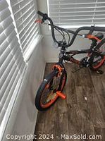 Children's black and orange bicycle shown standing indoors near a window with white blinds. Training wheels and handlebar details are visible.