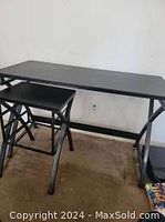 Photo showing the black computer table and matching black folding chair against a white wall with carpeted floor.