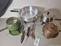 Four vintage fondue pots of different materials and colors with three burners underneath, and a collection of fondue forks with colored and wooden handles laid out with a fork holder.