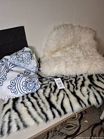 White fuzzy sheepskin pillow cover, faux zebra patterned fuzzy rug, and round mandala patterned fringed throw displayed together on a black surface
