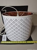 White metal hanging light with a perforated geometric pattern shown from the side with attached plug-in electrical cord and measuring tape below showing approximately 12 inches wide.
