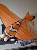 Full view of the large orange dory fish kite with distinctive stripes and eyes, coiled tail, kite line spool, and packaging.