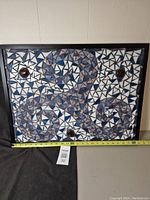 Front view of the large framed mosaic art piece showing blue, white, and purple triangular glass pieces arranged in swirls with three embedded glass accents and black frame. A measuring tape at the base shows size.