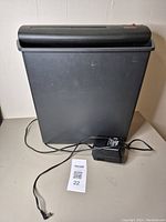 Front view of black plastic electric paper shredder, showing power adapter and cable.