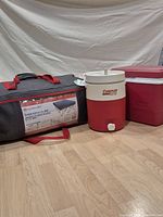 Full view of the lot showing the Outbound queen size inflatable bed in its folded carrying bag with red handles, the Coleman PolyLite2 drink cooler with white spout, and the red Rubbermaid personal cooler with white lid.