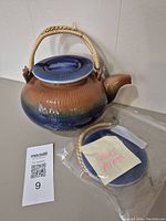Photo of the brown and blue glazed tea pot with a woven handle and a blue lid on top. Extra lid and handle parts visible in plastic packaging beside it.