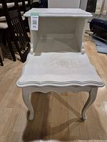Front view of white French provincial style step side table showing scalloped edges, curved legs, and upper and lower stepped surfaces with visible wear on paint finish.