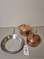 Set of two copper pots with lids and a stainless steel frying pan with brass handles placed on a gray surface.