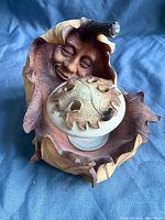 Candle holder shaped like an oak leaf with a smiling anthropomorphic tree face, removable leaf lid, and glass candle insert inside, resting on a blue fabric background.