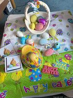 Photo of assorted Easter decorations including plastic tablecloths with eggs and chicks, woven basket with plush bunny and chick, various colorful Easter eggs, a yellow chick hat decoration, and Easter themed cake toppers.