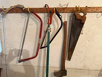 Photo showing set of various hand tools hanging on wall-mounted wooden rack including two bow saws, one pole saw, one axe, and one sickle.
