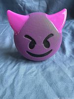 Front view of purple Bluetooth speaker shaped like devil face emoji with pink horns, showing facial features on speaker grill.