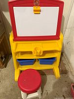 Front view of Step2 Flip and Doodle easel with white dry-erase board and blue storage bins, yellow base, with stool in front.