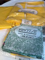 Photo of three oblong tablecloths, two vinyl in original wrap with brand 'Vinyle' and sizes 60" x 84", one yellow fabric tablecloth.