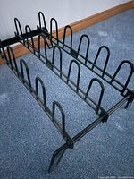 Black metal boot rack with 9 slots for holding pairs of footwear on carpeted floor near wooden baseboard.
