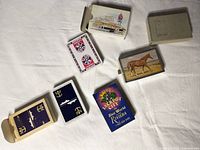 Four packs of vintage playing cards, two with illustrated cardboard boxes and two showing card back designs, all laid out on white surface.