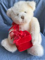 Photo of a vintage cream-colored Gund teddy bear sitting and holding a red velvet gift box tied with a red satin bow around its neck.
