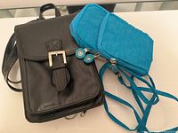 Photo shows two travel purses side by side on a glass surface: one black with a flap and metal buckle, and one turquoise with multiple zippers and long straps.