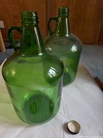 Photo showing two green glass wine carboys with handles on a floor surface, a metal cap lying in front of one.