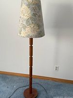Full view of the wooden standing lamp showing segmented wooden pole and floral fabric shade against plain wall and carpet flooring.