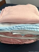 Stack of three folded wool blankets in pink, light blue, and beige colors on a bed, with a pink zippered storage bag partially visible under the blankets.