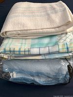 Stack of three wool blankets. Top blanket white with light pastel stripes, middle a multi-colored plaid, bottom solid light blue inside a plastic bag.