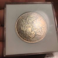 Front view of the 1974 British Commonwealth Games silver coin within a clear plastic case, showing detailed athlete figures and text.