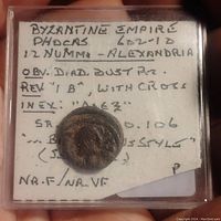 Front view of the ancient bronze coin inside a protective case with handwritten label showing details like Byzantine Empire Phocas 602-610 AD, 12 Nummi Alexandria, obverse with diademed bust, reverse with cross, and condition notes.