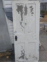 Solid hardwood antique door standing upright adjacent to a white wall with paint chipping off exposing underlying layers and wood. Shows the door handle and lock hardware on one side.
