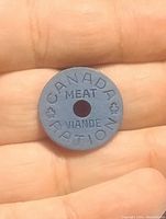 Top view of blue Canadian meat ration token held in hand showing front face with text 'CANADA MEAT VIANDE RATION' and central hole.