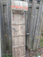Front view of a vintage large wooden toboggan sled leaning against a wooden fence showing the vertical wooden slats, metal runners, attached rope, and faded red paint on the curved front.