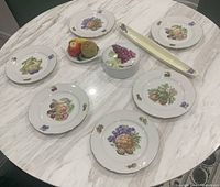 Top view of six JL Menau fruit plates arranged around ceramic lidded dish, ceramic fruit basket decoration with two ceramic fruits, and long grape dish on marble table.