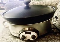 Oval Crock-Pot slow cooker with lid on, showing stainless steel body and control panel with two knobs and digital timer.