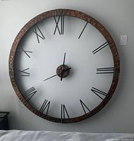 Front view of large metal wall clock hung on a white wall showing Roman numerals and black clock hands.