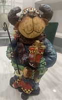 Front and slightly angled view of a wooden-style moose statue wearing festive clothing, holding a red gift and staff.