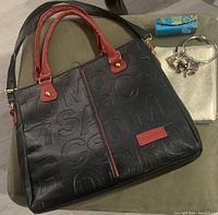 Black handbag with red handles and embossed lettering, displayed with small wallet, card holder, and charm bracelet