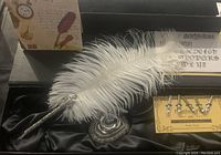 Top view of calligraphy kit including feather pen with silver decorative handle, nibs set, ornate pen holder, all on black satin inside box, with outer carton featuring calligraphy illustrations.