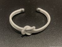 Photo showing front side of silver plated mesh cuff bracelet with knot detail, placed on dark surface.
