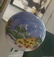 Photo of hand painted glass ornament showing winter village landscape with yellow houses and snow-covered trees under a blue sky.