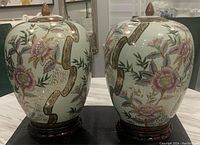 Pair of signed hand-painted Chinese ginger jars/urns with lids and wooden bases shown from front, decorated with floral and bamboo designs accented in gold.
