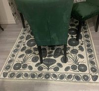 Photo showing off-white rug with large black floral pattern beneath green velvet chairs around a dining table.