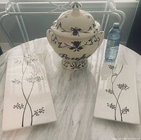 Photo showing large ceramic lidded soup tureen with dark blue floral patterns and two rectangular white ceramic trivets with black botanical designs, along with a water bottle for scale.