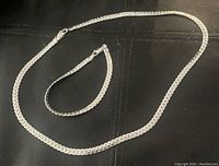 Top-down view of the necklace and bracelet laid flat on a black surface showing their silver finish and flat linked chain design.