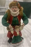 Front view of the vintage Santa Claus and child statuette, showcasing Santa in green and red attire with fur trim, holding a child in a green dress.