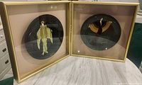 Two framed round plates with Art Deco hand painted female figures displayed side by side on a white surface under lighting. Plates are framed in gold shadow boxes with glass covers.