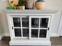 Front view of wooden cabinet with two sliding glass paneled doors in distressed white finish with decorative top molding and base.