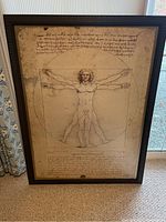 Full view of the framed Vitruvian Man print leaning against a window, showing the entire artwork and frame.