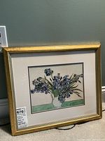 Framed print artwork of Vincent Van Gogh's 'Irises' leaning against wall, showing gold wooden frame and floral print.