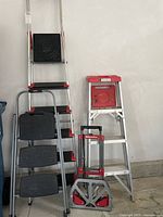 Photo of three ladders of different sizes and a small dolly, showing orange, red, black and silver colored ladders and dolly in garage.